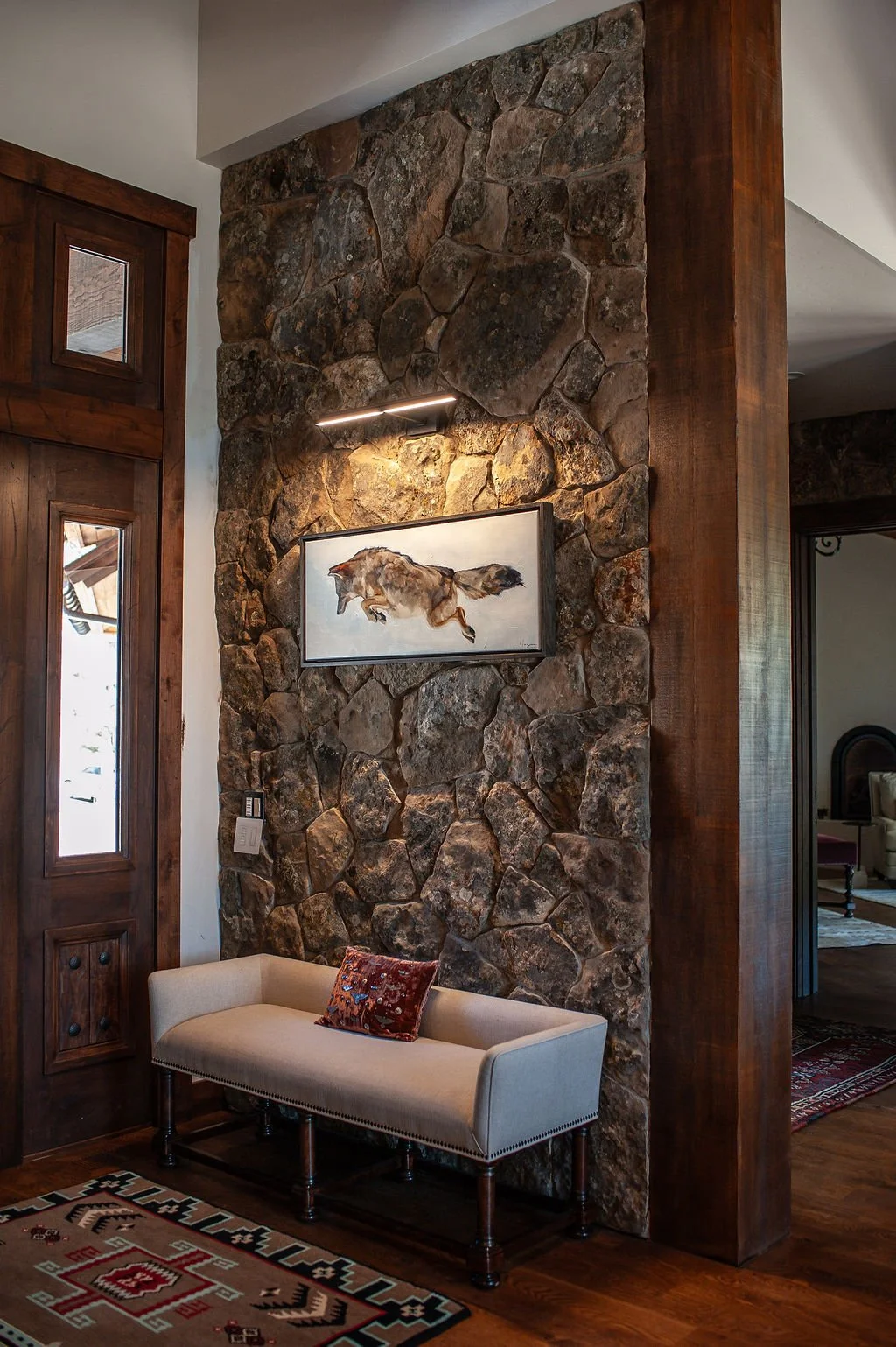 Interior of a room with a rustic stone wall, a framed painting of a running horse, a small wall-mounted light above the painting, a white upholstered bench with a small red decorative pillow, a wooden floor, and a large wooden frame door with glass p