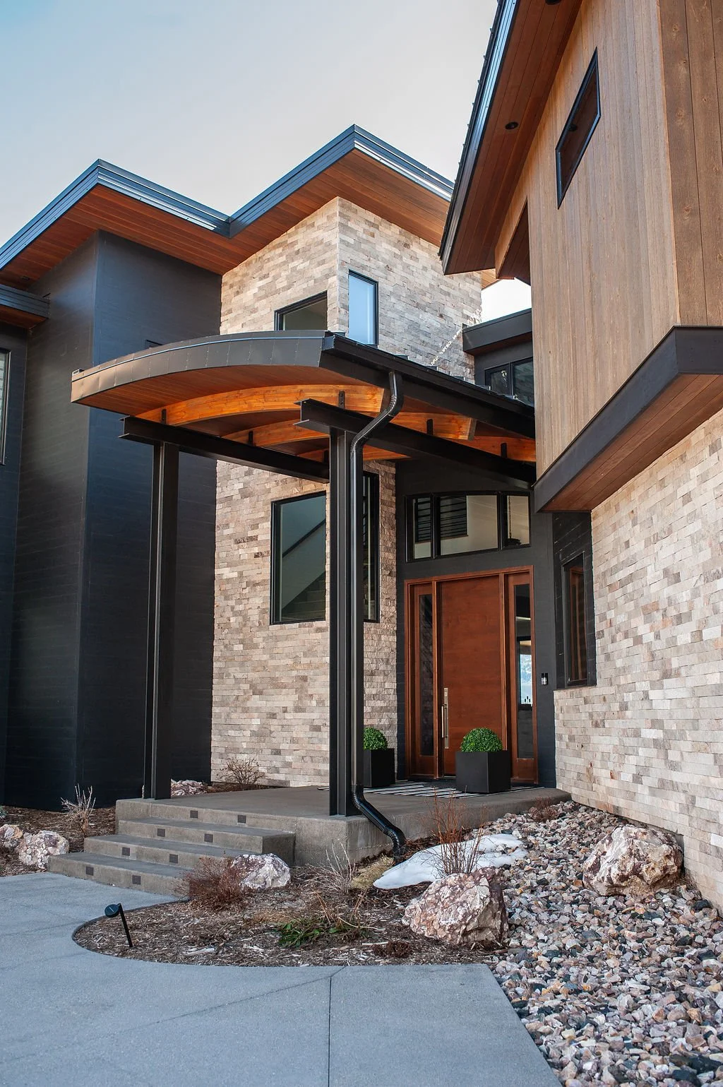 Modern house with brick, black wood siding, and wood accents, featuring a concrete staircase, large windows, and a landscaped front yard with rocks and plants.
