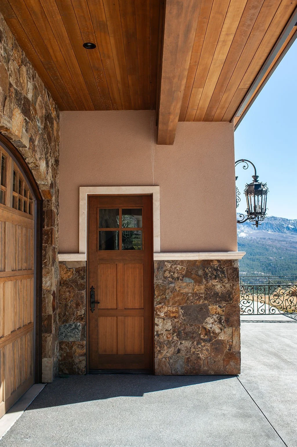 Exterior view of a house featuring a wooden door with a small window, a stone wall, a wooden ceiling, and an outdoor lantern, with a mountain landscape in the background.
