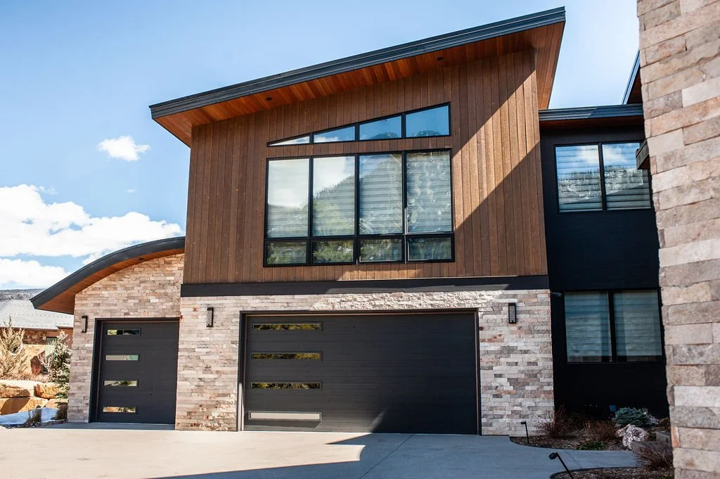 Modern two-story house with a brick and wood exterior, black garage doors, large windows, and a sloped roof under a partly cloudy sky.