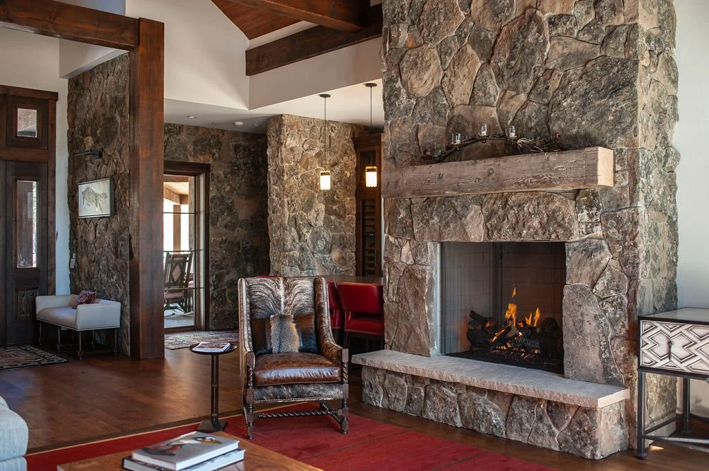 A cozy living room with a large stone fireplace, wooden beams, and furniture like an armchair, side table, and a white bench near a doorway, in a rustic style.