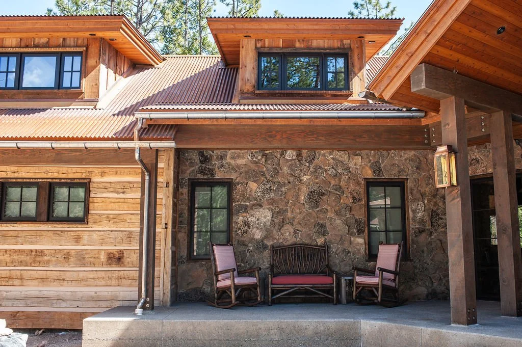 Close-up of a rustic house exterior with stone and wood siding, featuring a small covered patio with two chairs and a bench.