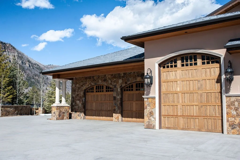 Photo of a house with three wooden garage doors, stone and stucco exterior, mountain landscape, and blue sky with clouds.