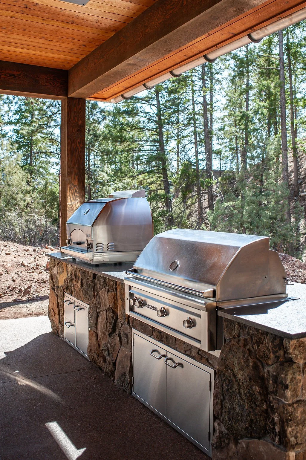 Outdoor outdoor kitchen area with two stainless steel grills on a stone counter under a wooden roof, surrounded by trees.