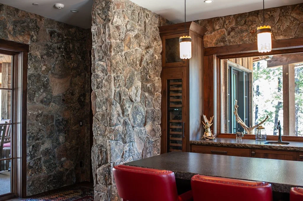 Interior of a rustic kitchen with stone walls, a large window with wood trim, and a granite countertop. Two hanging pendant lights and red chairs are visible.
