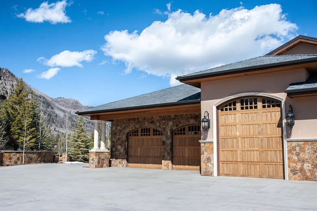 A house with a stone and stucco exterior, a dark gray roof, and wooden garage doors, situated near mountains with trees under a partly cloudy sky.
