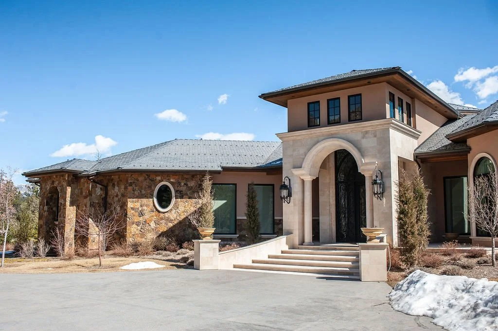 Large upscale home with stone and stucco exterior, arched entrance, steps leading up to door, and landscaping, under a blue sky with some clouds.