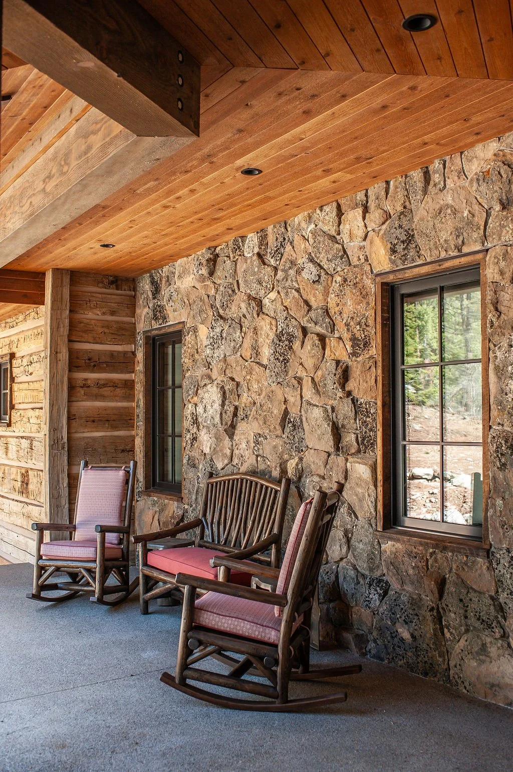 Porch area with rustic stone wall and wooden ceiling, featuring two wooden rocking chairs with pink cushions, and a window showing outdoor greenery.