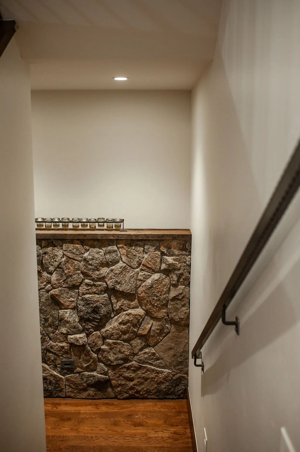 A staircase with a stone wall on the left side, a wooden floor, and a metal handrail on the right side, leading to a small landing with a row of candles on a ledge.