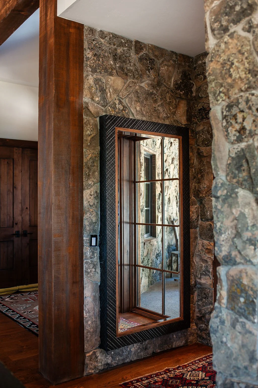 A rustic interior with stone walls, wooden beams, and a large mirror with a black frame reflecting a stone wall and a window.