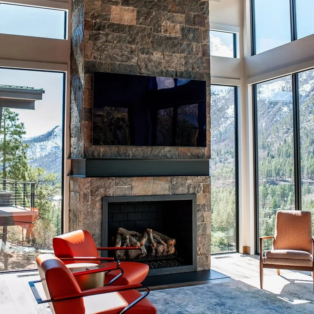 Living room with a mountain view, large window, stone fireplace with a TV mounted above it, orange armchair, beige armchair, and wooden flooring.