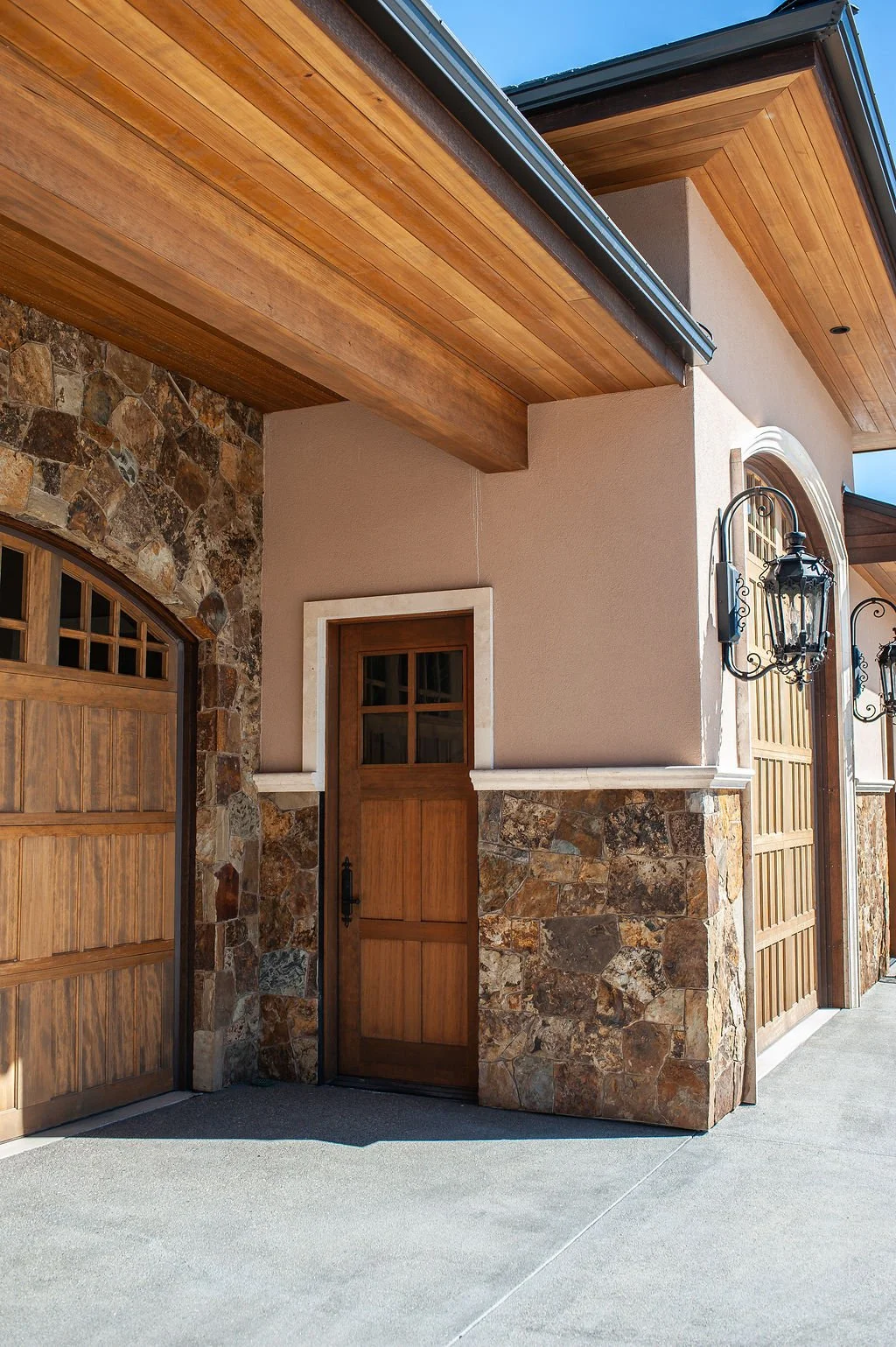 Exterior of a building showing a wooden garage door, a small wooden door with a window, stone and stucco walls, and decorative black iron lanterns.