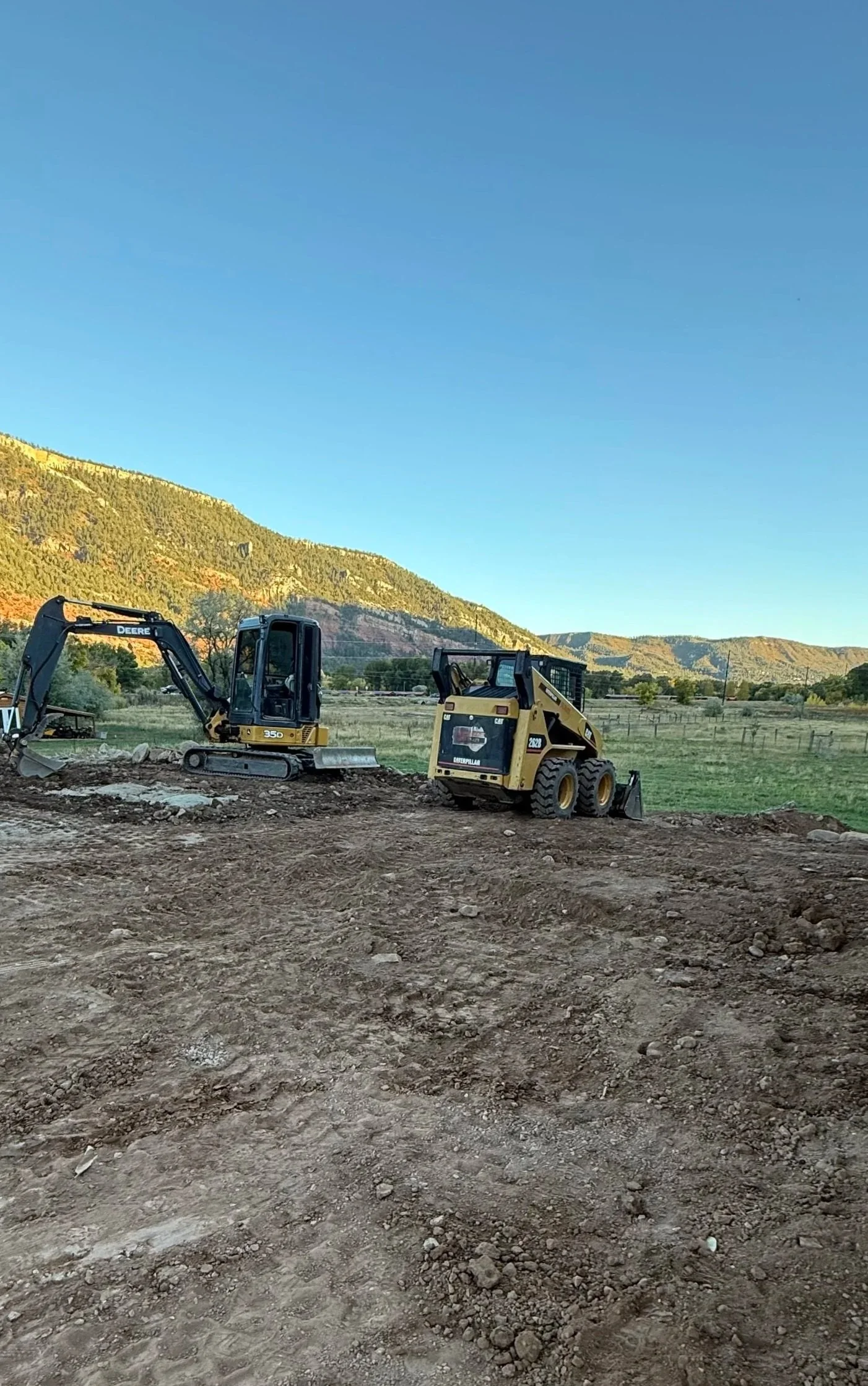 Two construction vehicles, a small excavator and a skid steer loader, working on a dirt area with mountains and clear blue sky in the background.