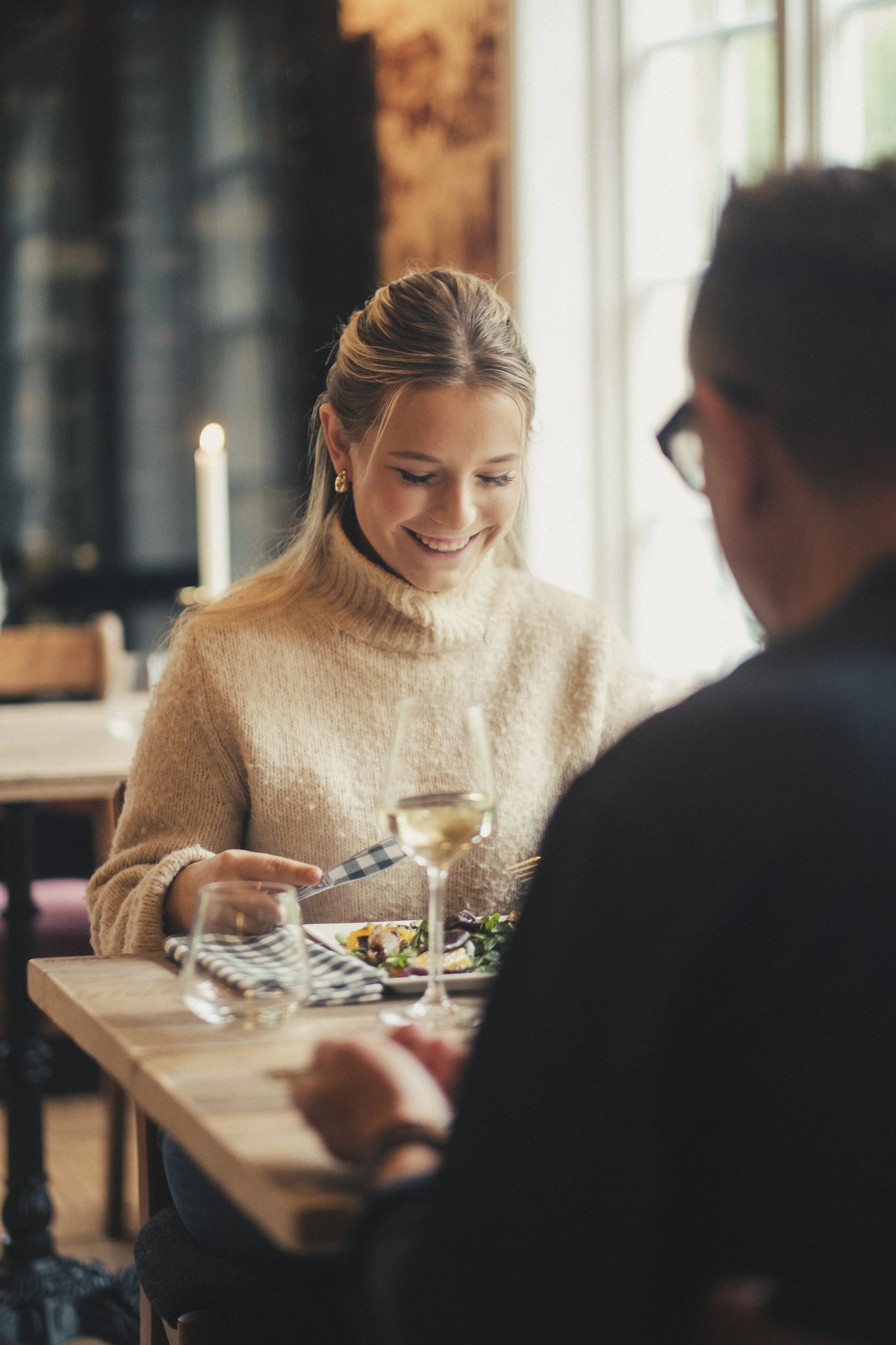 En kvinne smiler mens hun spiser på Bygdøy Vertshus restaurant, med vin og mat på bordet.