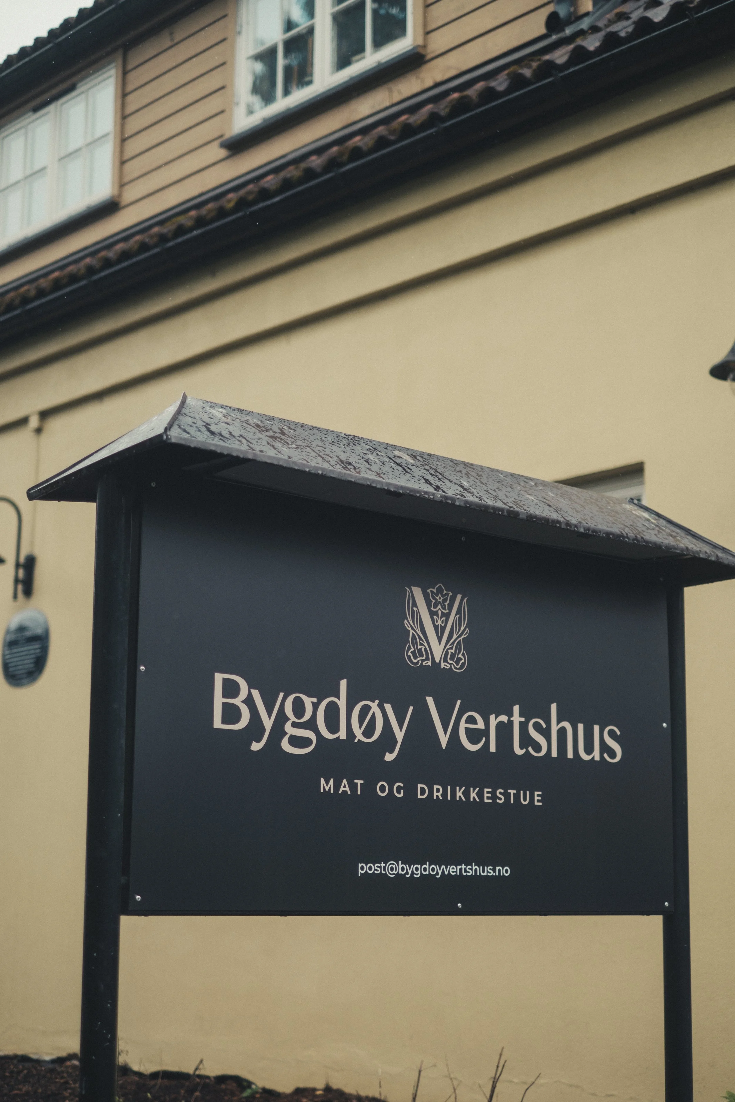 Et skilt med teksten «Bygdøy Vertshus» og kontaktinformasjon, plassert foran en bygning med lys gul vegg og tak av utseende takstein. Har et lite takkantbeslag over skiltet.