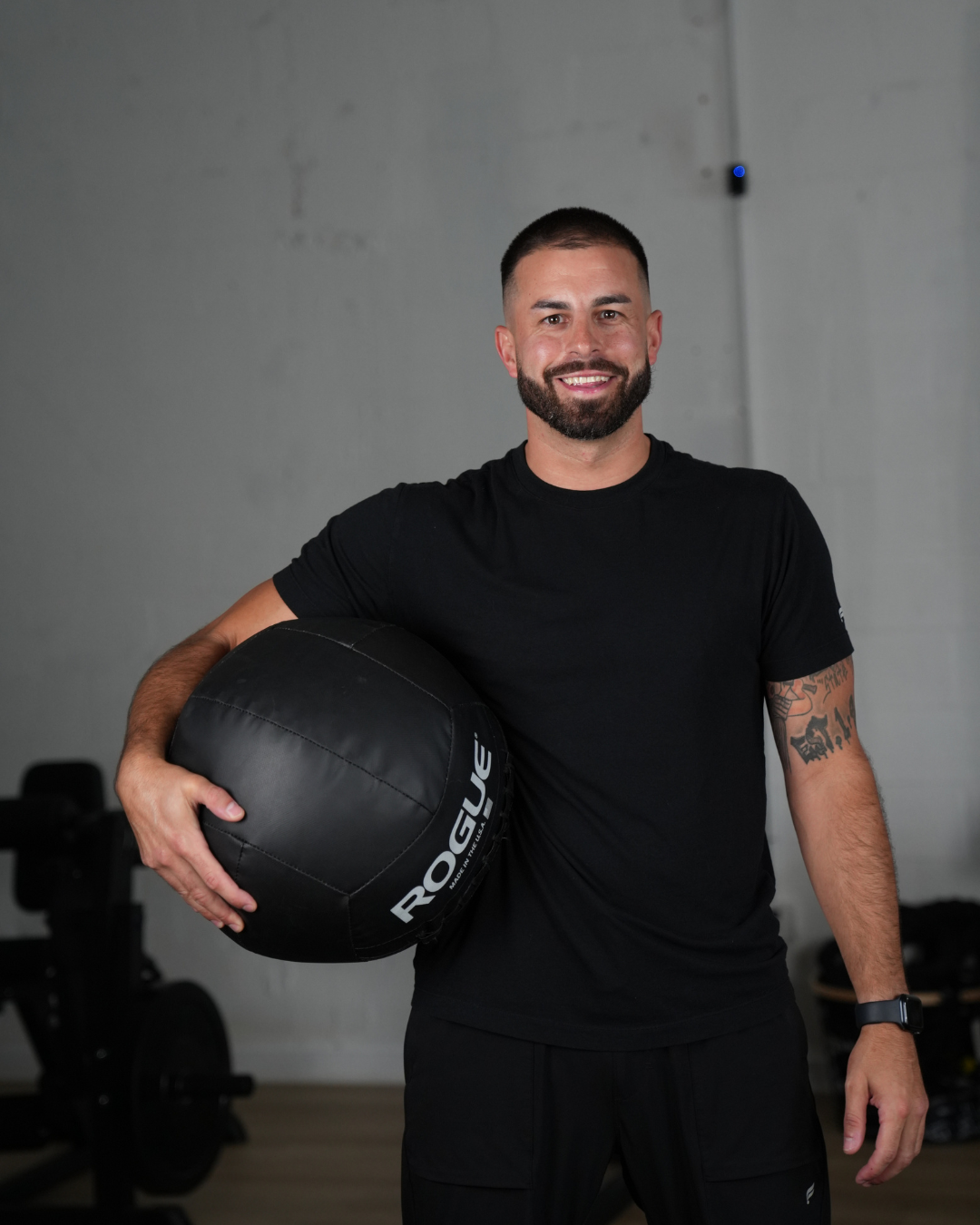 A smiling fit man with a beard and tattoos on his arm, dressed in black athletic clothing, holding a Rogue medicine ball in a gym setting.