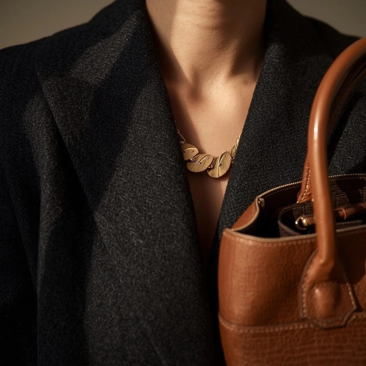 Close-up of a woman wearing a gold leaf-shaped necklace, a black textured blazer, and carrying a brown leather handbag.