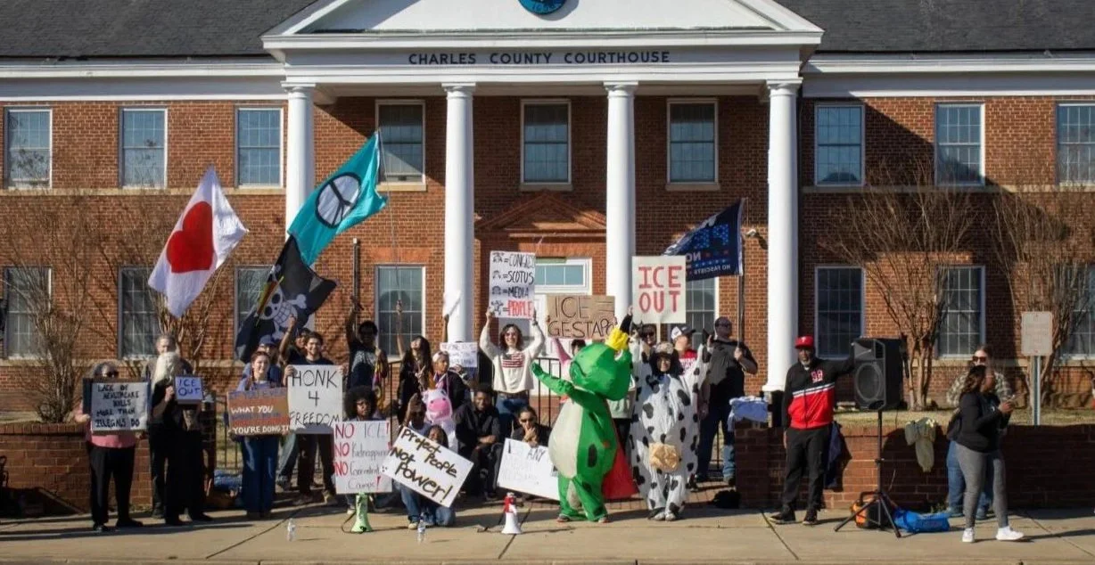 Protest outside Charles County Courthouse with people holding signs and flags, including a large green dinosaur costume, and a Cow costume.