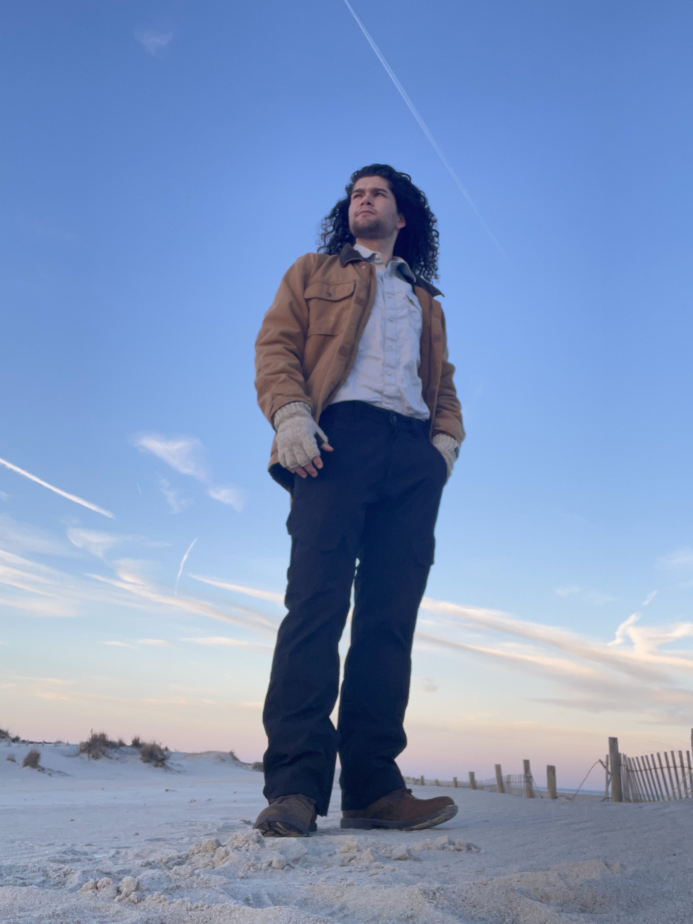 Evan Smith stands on Assateague National Seashore under a clear blue sky with a few clouds and contrails. He is wearing a Carhartt work jacket, white shirt, black pants, and brown shoes, and looks off into the distance.