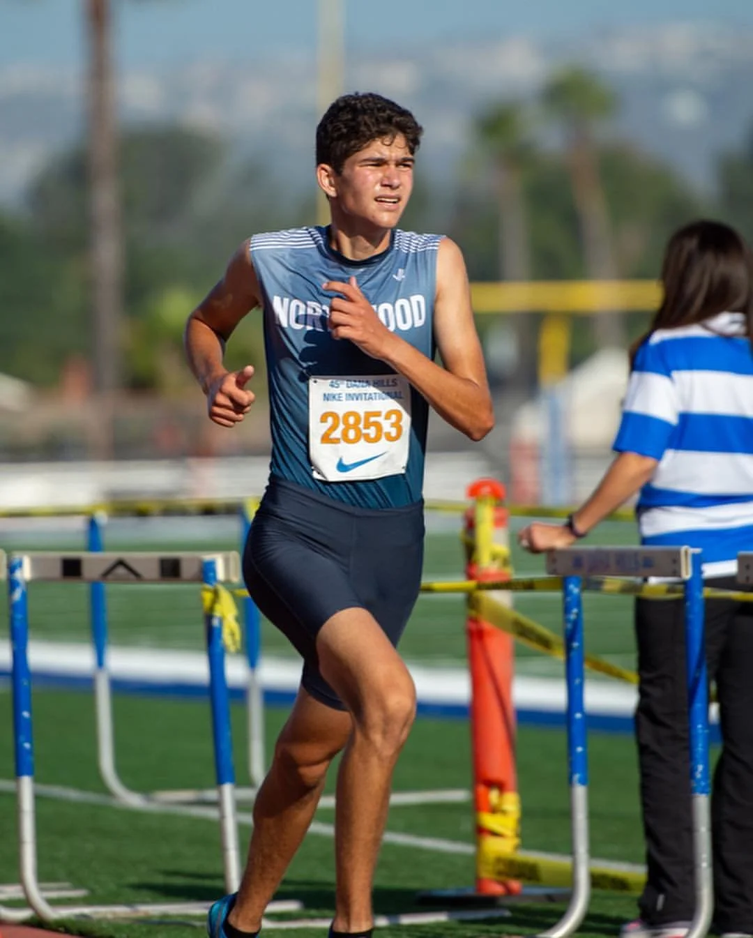 Evan Smith running a 1600m track race, wearing a blue and black athletic uniform during a sunny day. He appears focused and determined.
