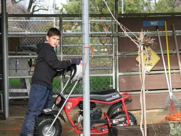 Evan Smith as a young boy wearing a black hoodie and blue jeans standing next to a red electric mini bike, under the roof of a stable with trees and a horse crossing sign in the background.