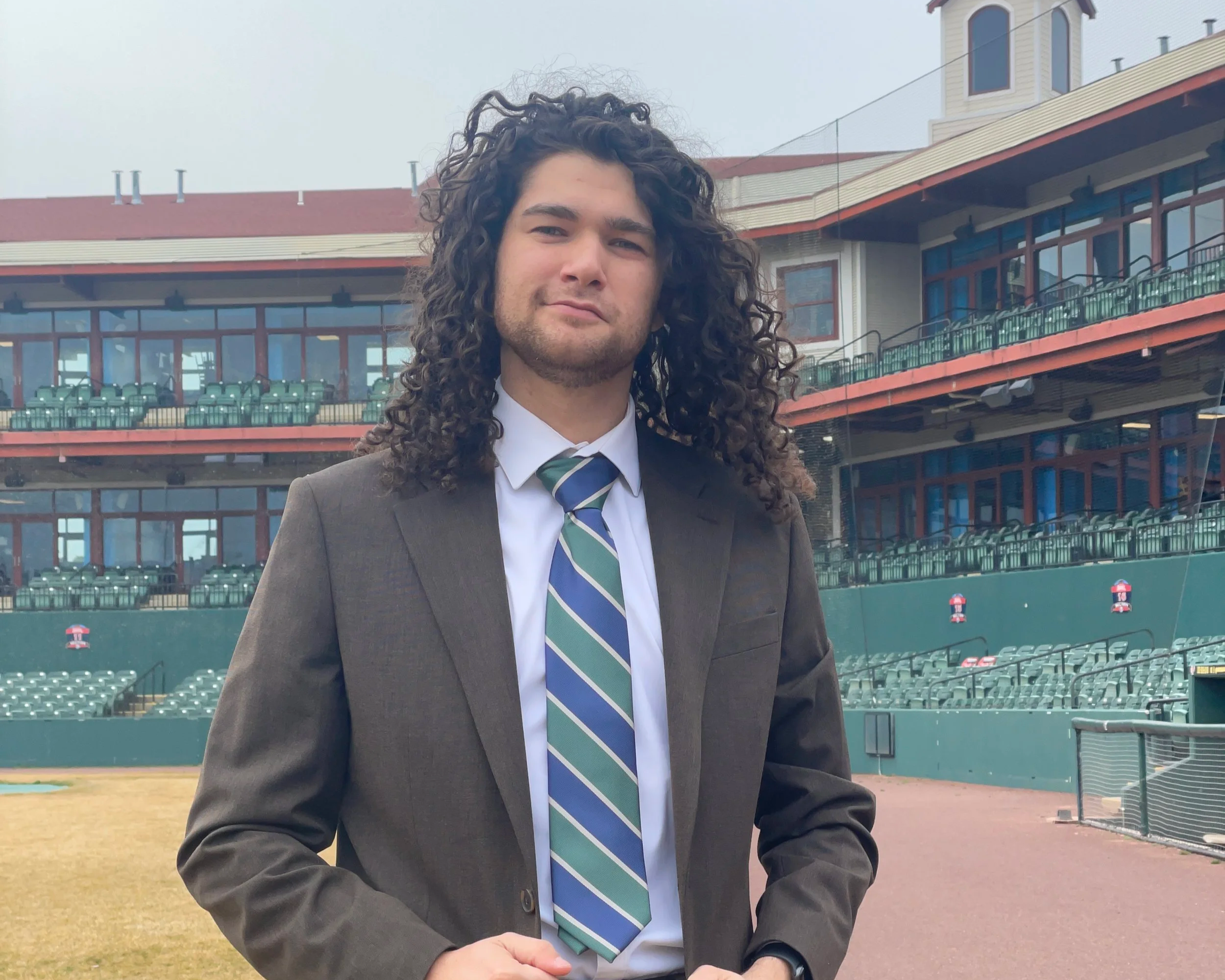 Evan Smith wearing a suit and striped tie, standing on the Blue Crabs baseball field with the stands in the background.