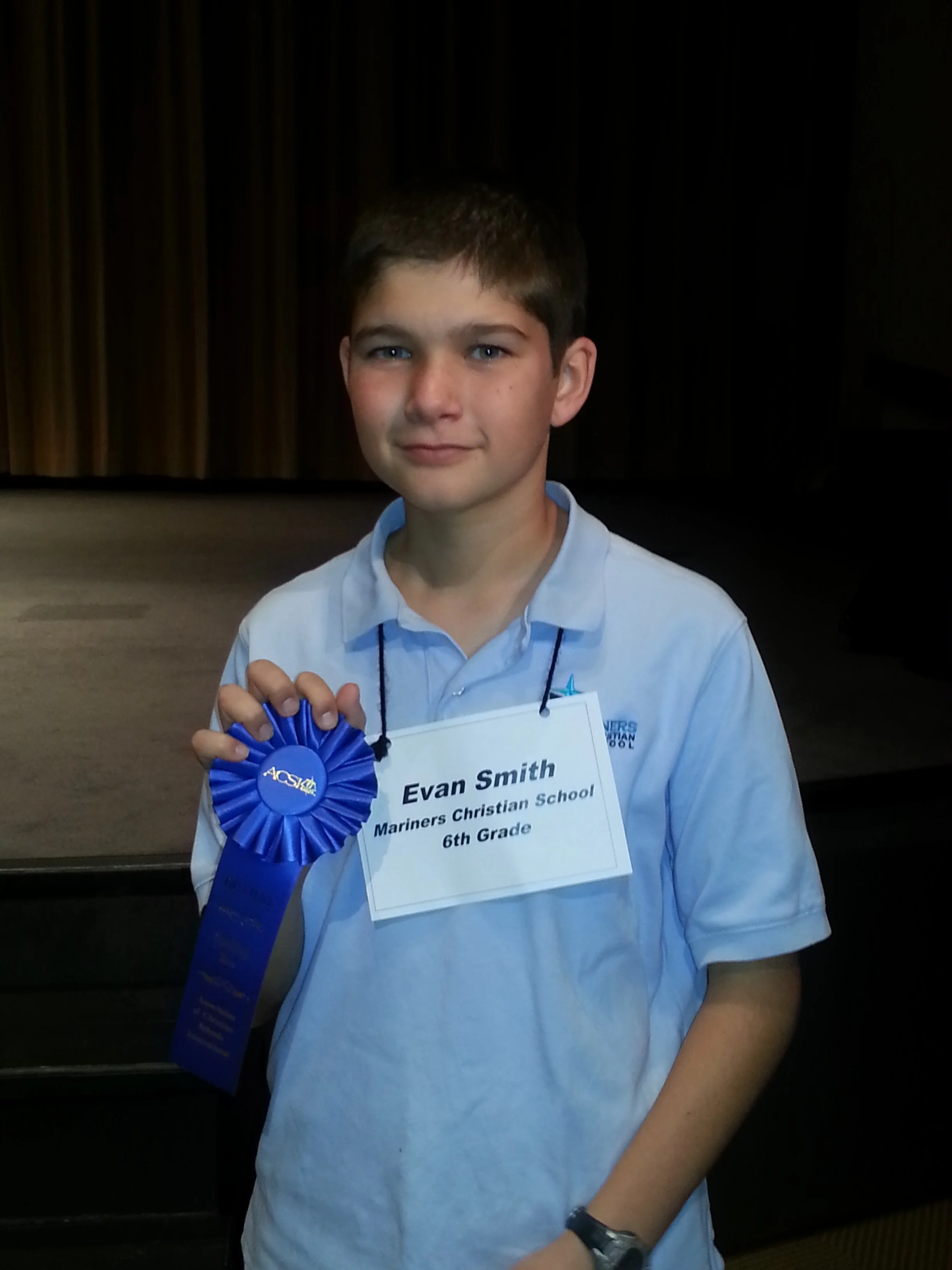 Evan Smith holding a blue ribbon award for the spelling bee, with a name tag that clarifies he is in the 6th grade.