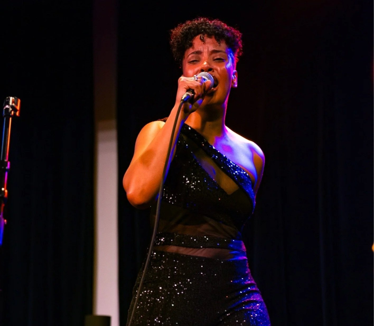 A woman singing into a microphone on stage, wearing a black sparkly dress, with dark background and stage lighting.