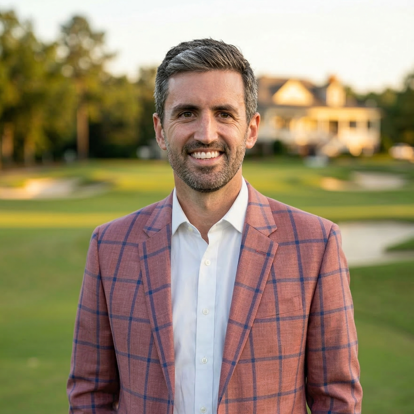 Portrait of a smiling man in a pink plaid blazer and white shirt outdoors on a sunny day, with a house and trees in the background.