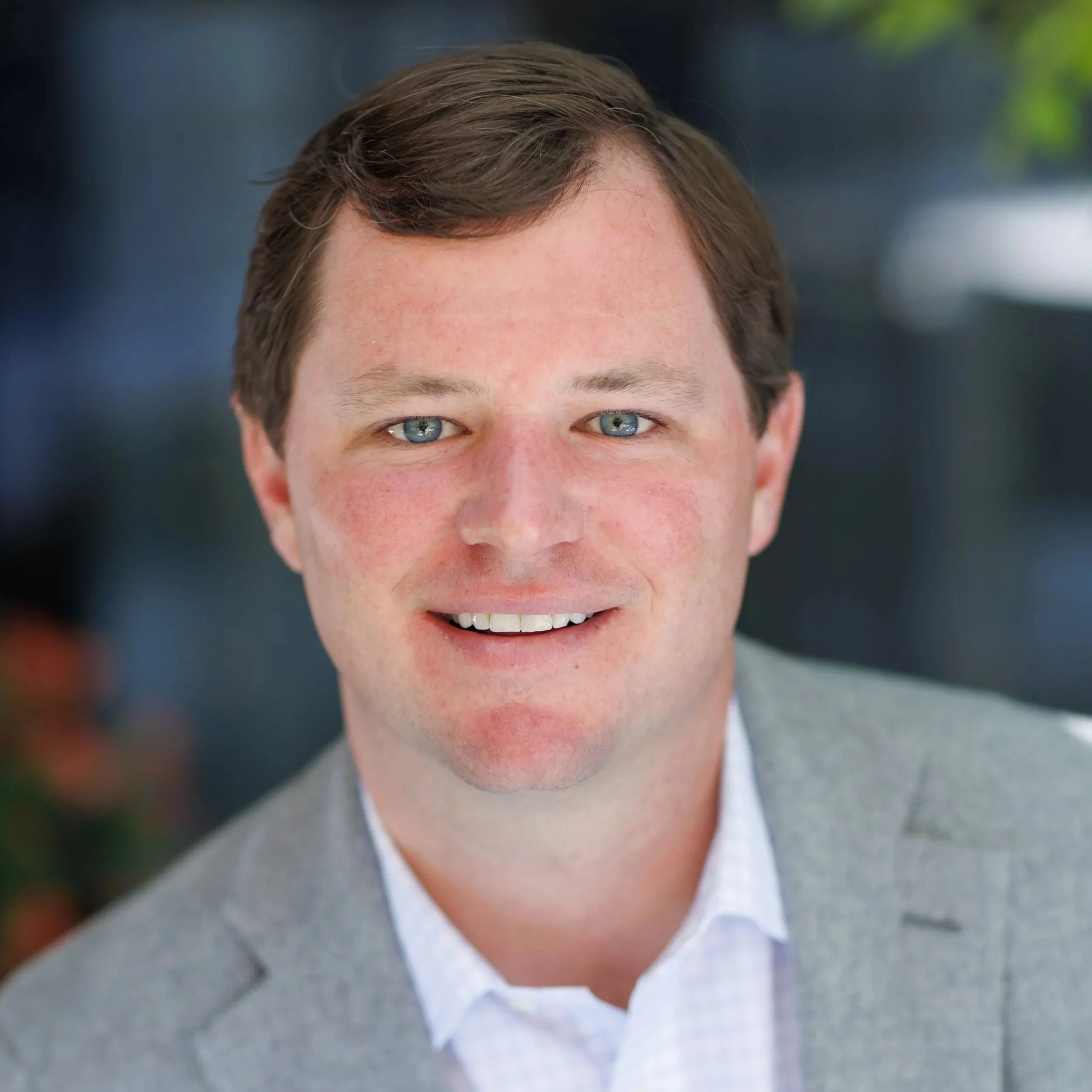Close-up portrait of a smiling man with blue eyes and brown hair, wearing a gray blazer and white shirt, outdoors with a blurred background.