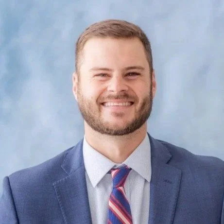 A smiling man in a blue suit with a tie, standing against a soft blue background.