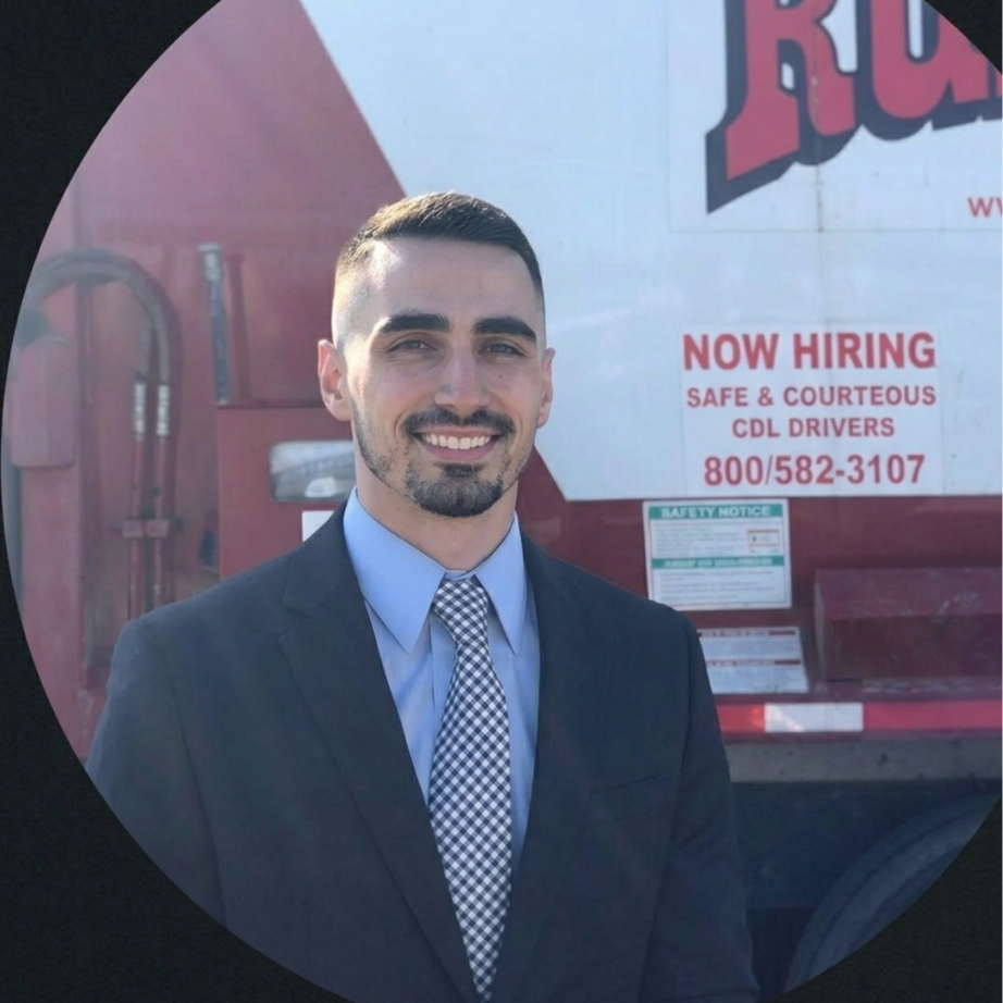 A man in a suit with a blue shirt and checkered tie smiling in front of a red truck with job hiring signs.