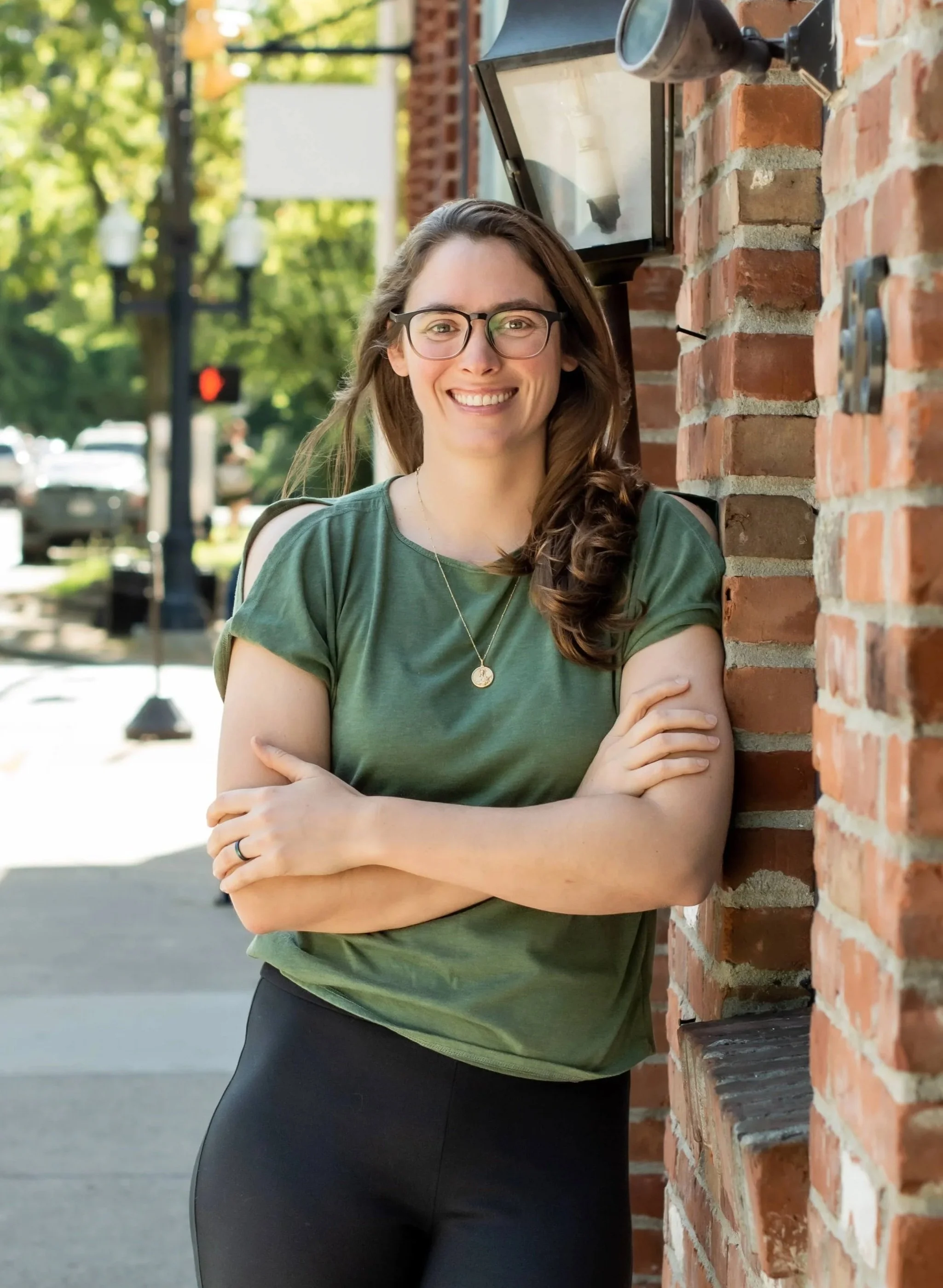 A young woman with glasses, long brown hair, and a green t-shirt standing outside against a brick wall, smiling with arms crossed.