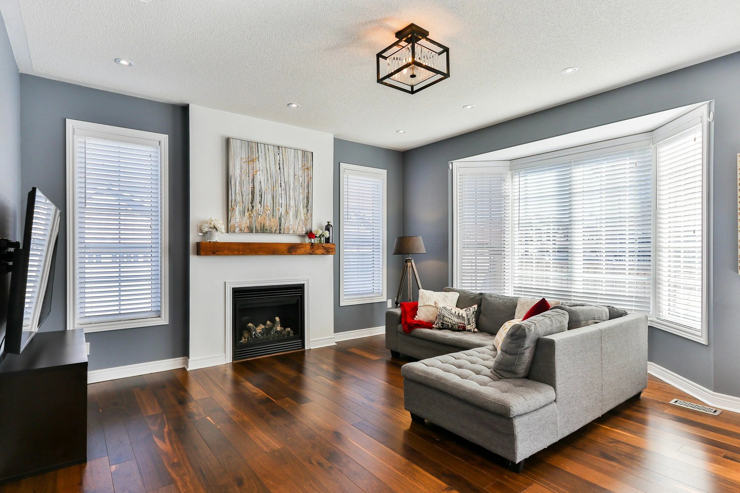 Living room with gray walls, hardwood floors, a fireplace with a wooden mantle, a gray sectional sofa with decorative pillows, large bay windows with white blinds, a floor lamp, and a ceiling light fixture.