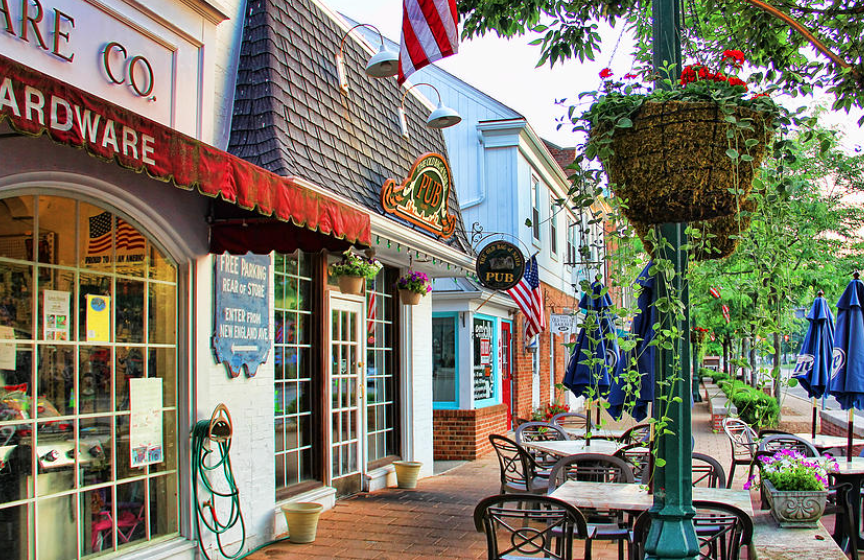 Empty outdoor patio of a pub in a small town, with tables, chairs, blue umbrellas, hanging flower baskets, and American flags. Stores with colorful storefronts line the sidewalk.