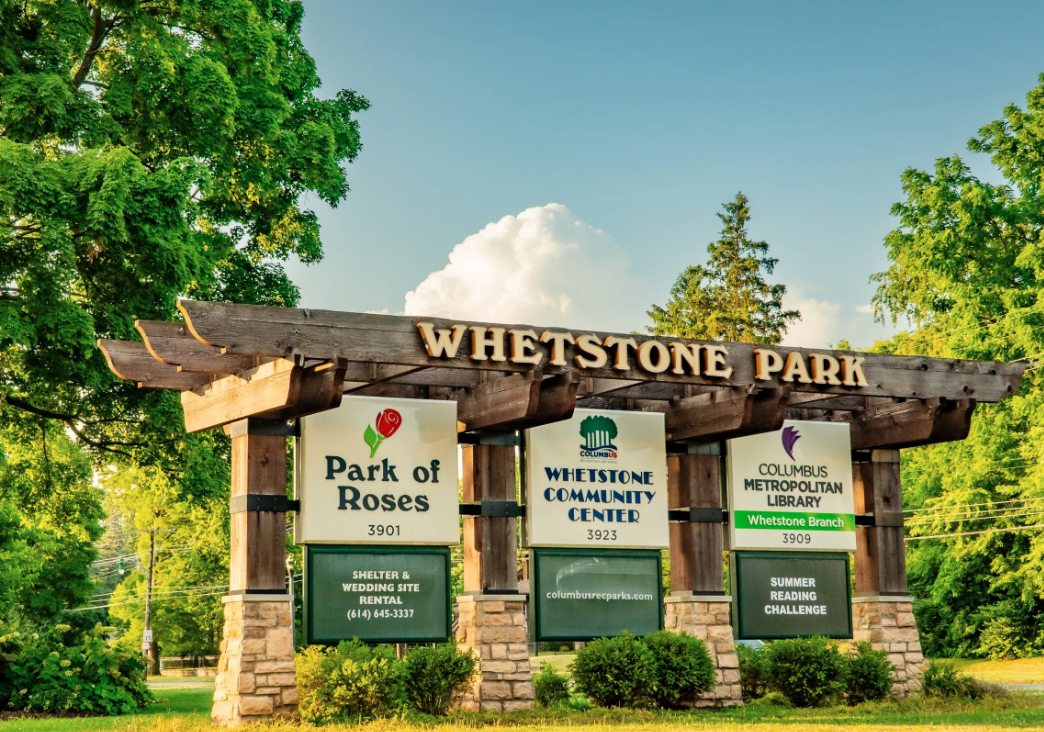 Signboard at Whetstone Park listing Park of Roses, Whetstone Community Center, and Columbus Metropolitan Library Whetstone Branch with green trees and blue sky in the background.