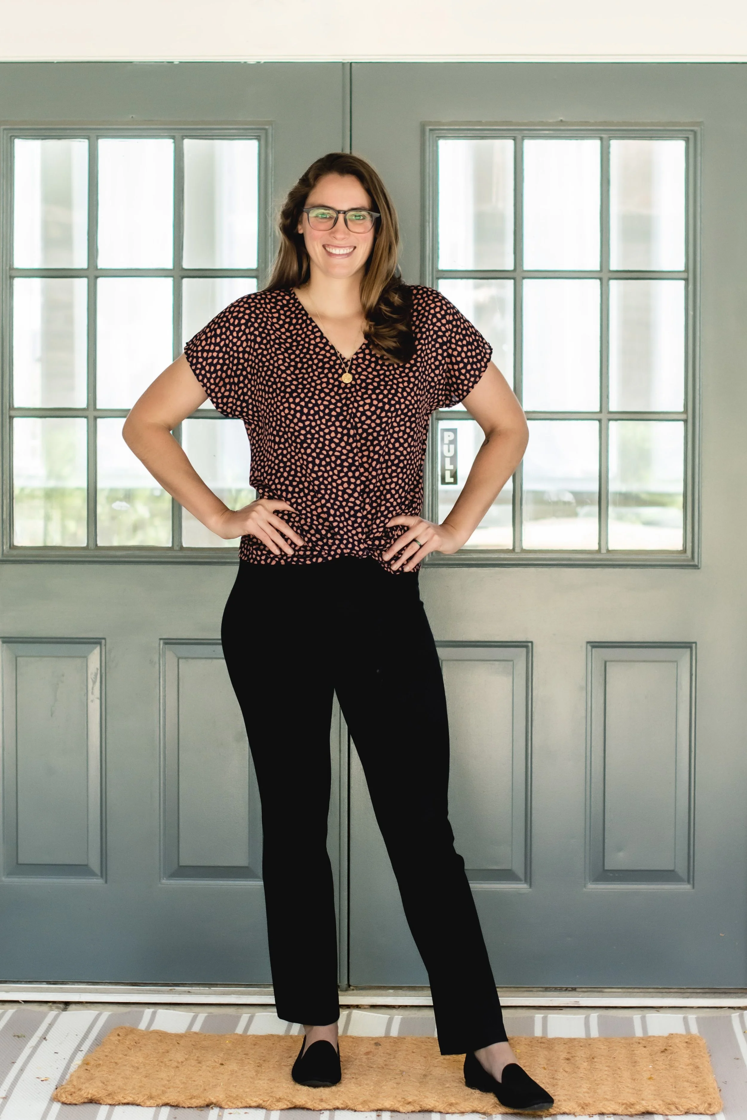 A woman with glasses and long brown hair standing in front of double-pane glass doors with a 'PULL' sign, smiling and posing with hands on her hips, wearing a black and pink patterned blouse, black pants, and black slip-on shoes.