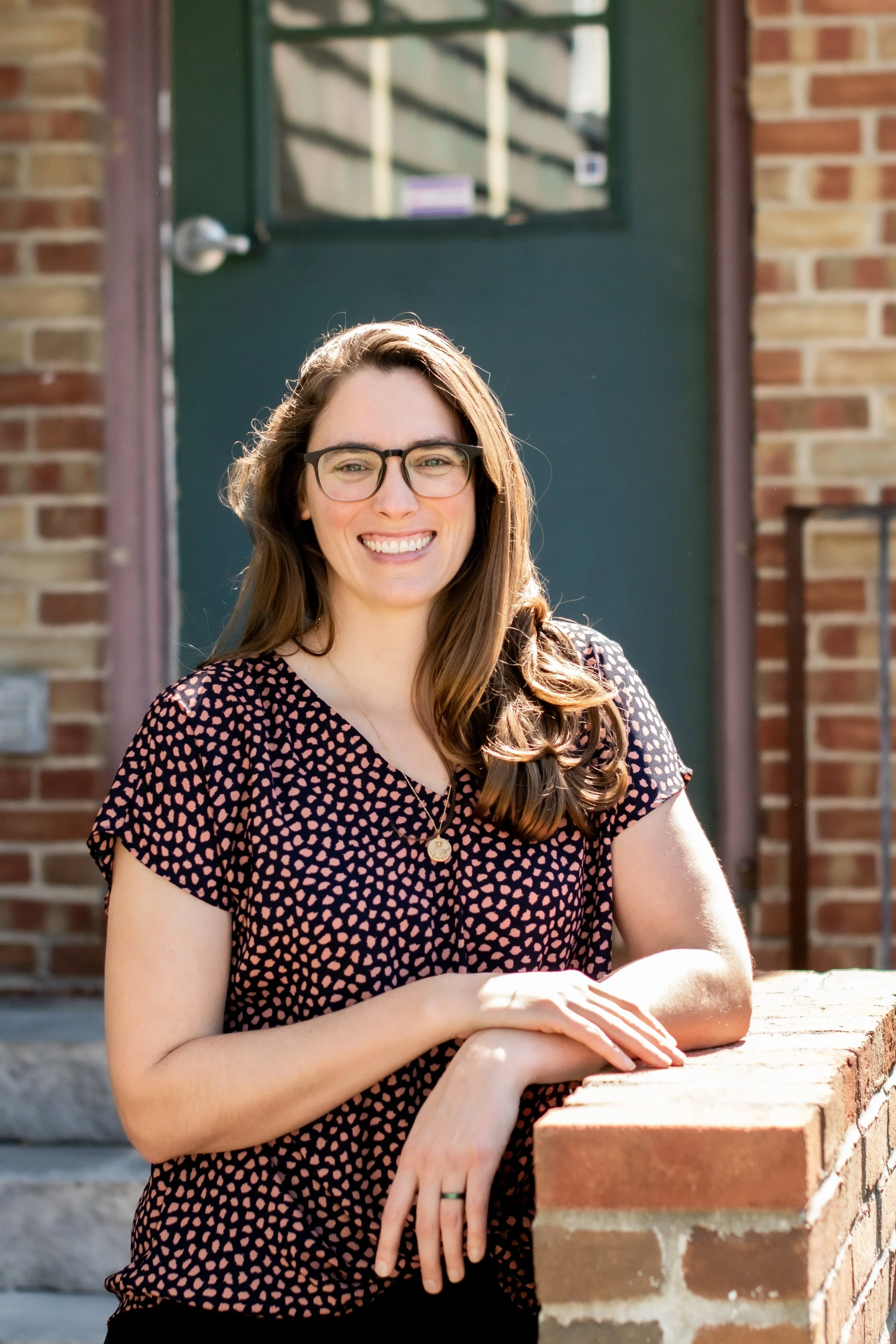 A woman with long brown hair, glasses, and a patterned blouse, smiling outdoors in front of a brick building and a green door.