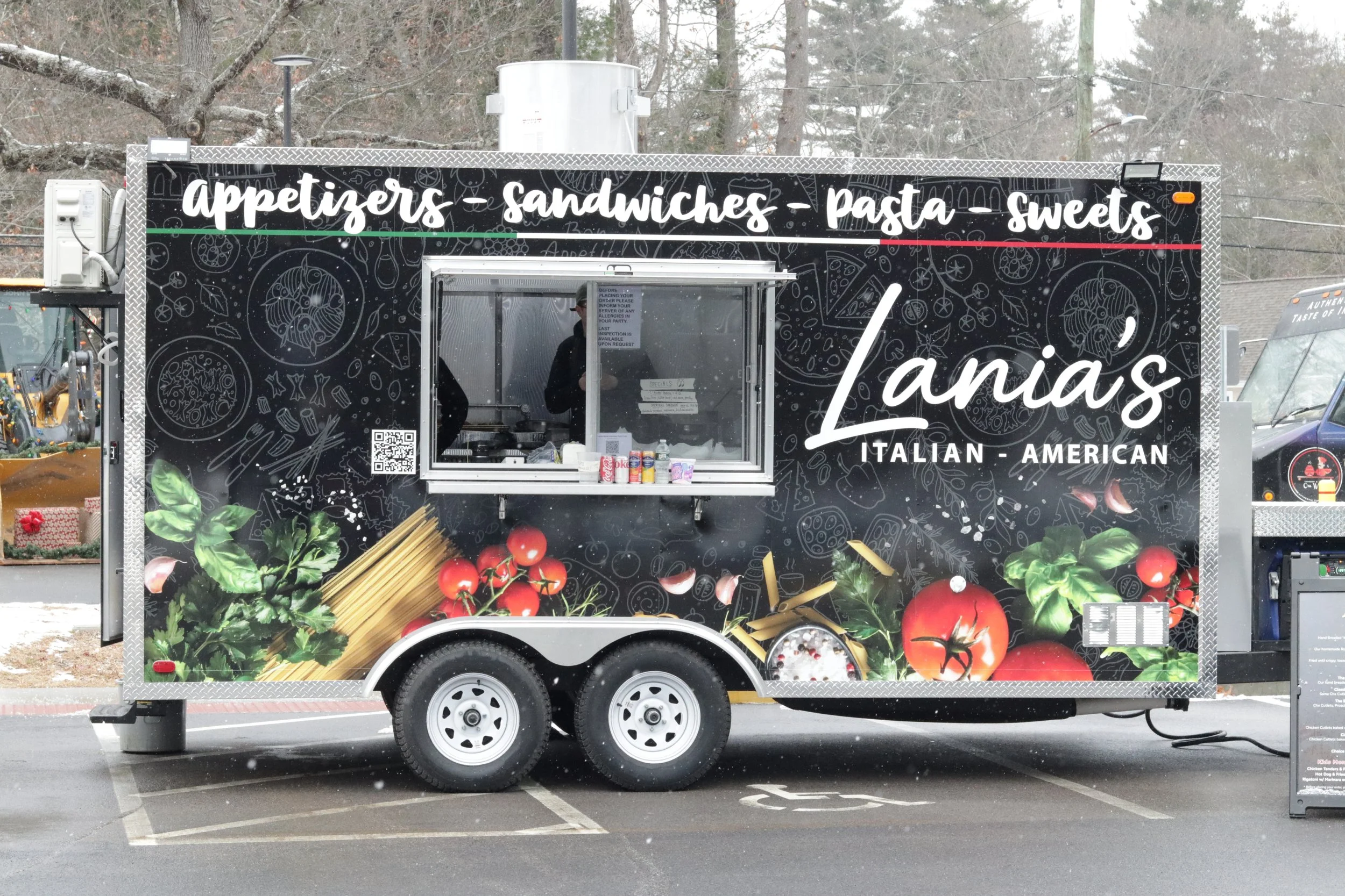 Food truck with a black sign displaying white text that reads 'appetizers, sandwiches, pasta, sweets, Lanas' and smaller text 'ITALIAN - AMERICAN'. The truck has a window with a person inside, and decorative images of vegetables and food items on the exterior.