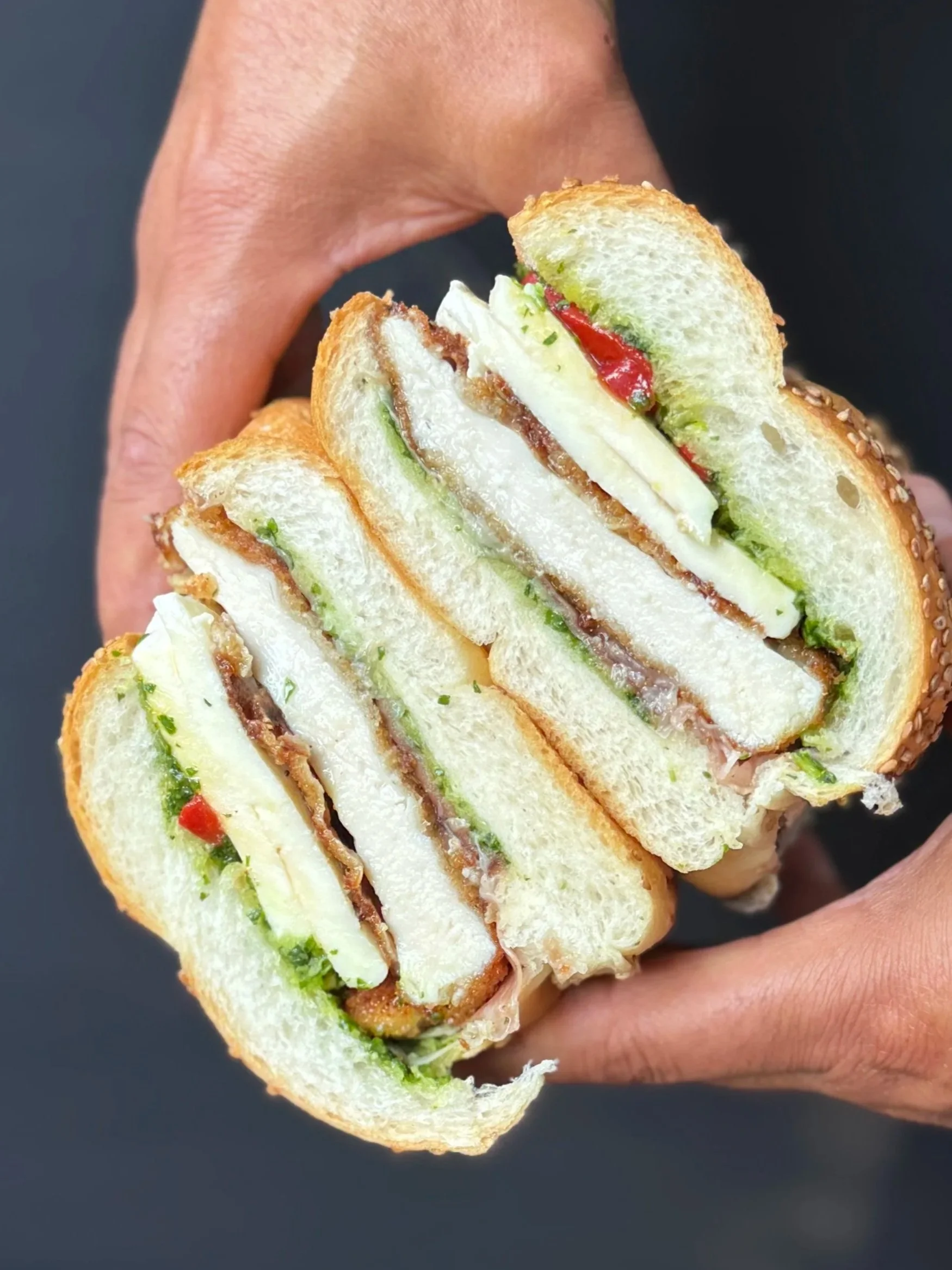 Close-up of a hand holding a cut sandwich with layers of lettuce, white cheese, a slice of turkey, tomato, and green peppers on a sesame seed bun against a dark background.