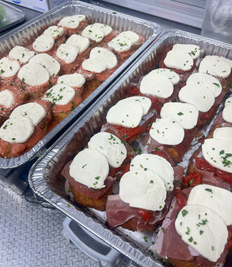 Two aluminum trays of prepared appetizers topped with mozzarella and chopped herbs, serving as finger foods or party snacks.