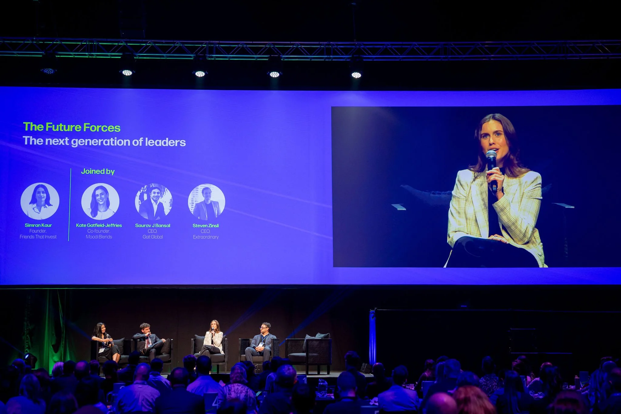 A large presentation screen displays a slide titled "The Future Forces: The next generation of leaders" with photos and names of four speakers, and a panel discussion with four people seated on stage, including a woman speaking into a microphone.