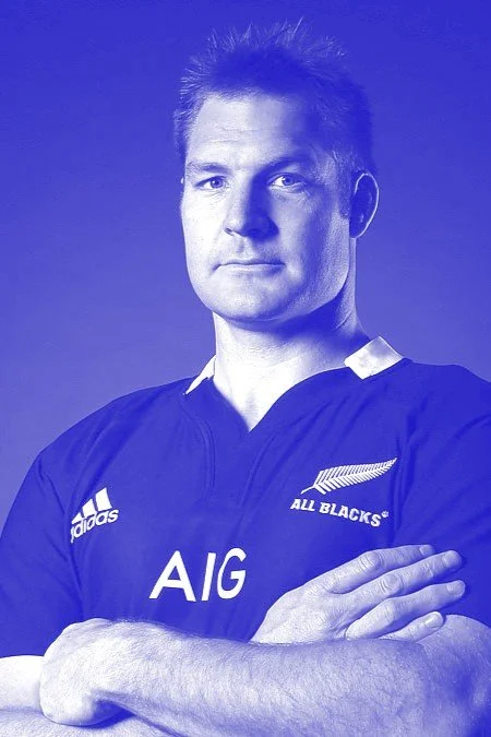 A man in a black sports jersey with the New Zealand All Blacks logo and the text AIG, standing with crossed arms against a blue background.