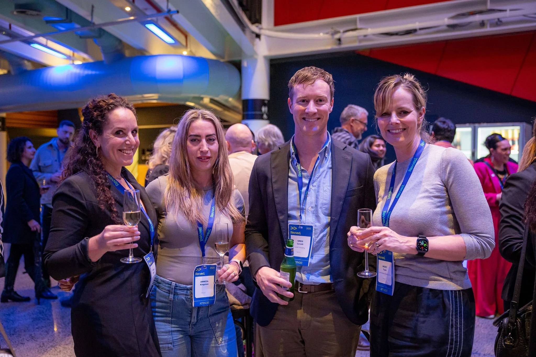 Group of four people smiling and holding drinks at a social or networking event with other attendees in the background.