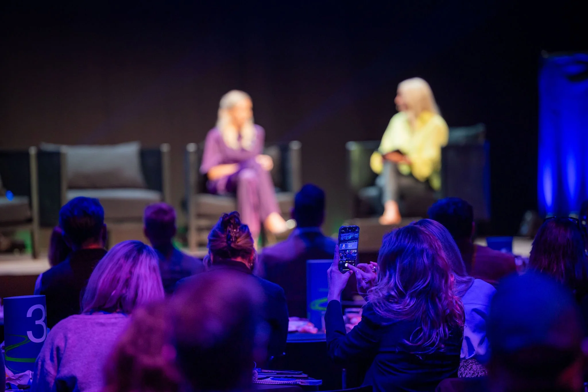 Two women are sitting on stage engaged in a discussion, with an audience watching. One woman wears a purple outfit, and the other wears a yellow top. The audience members are taking photos or videos, and there are numbered signs on the tables.