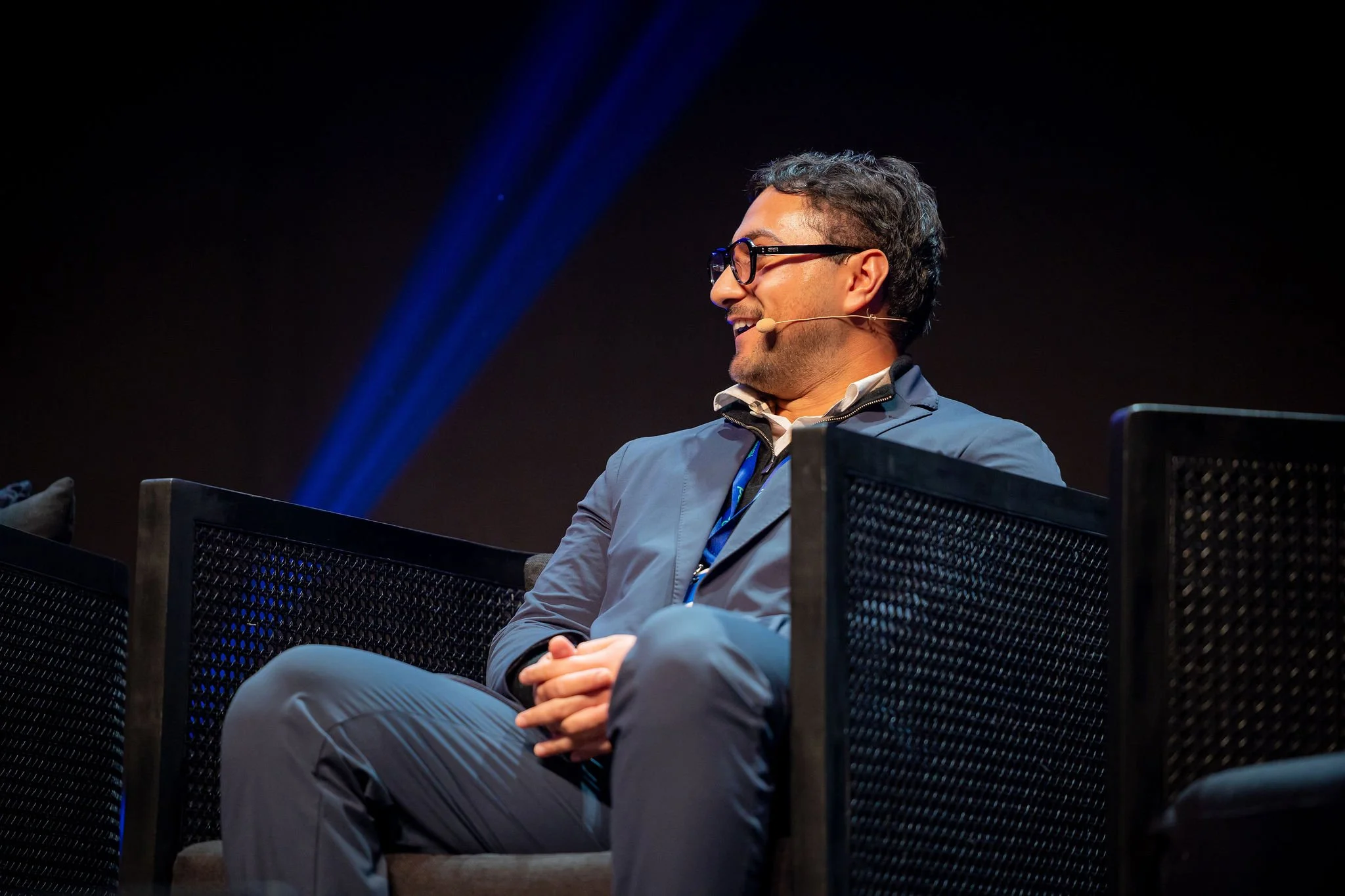Side view of a man with glasses, dark hair, beard, wearing a blue blazer, and white shirt, smiling while sitting on a black mesh chair on a stage with a dark background.