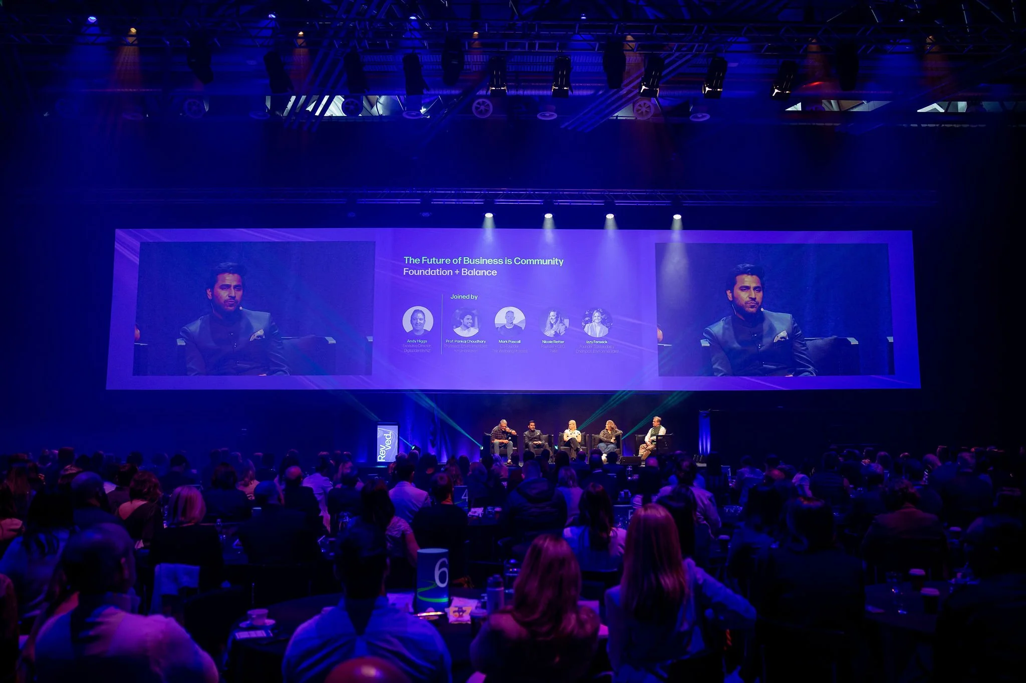 Revved panel discussion event in a darkened auditorium with stage lights. There is a big screen displaying a presentation titled 'The Future of Business is Community'. The audience is seated at tables, watching the panel on stage.