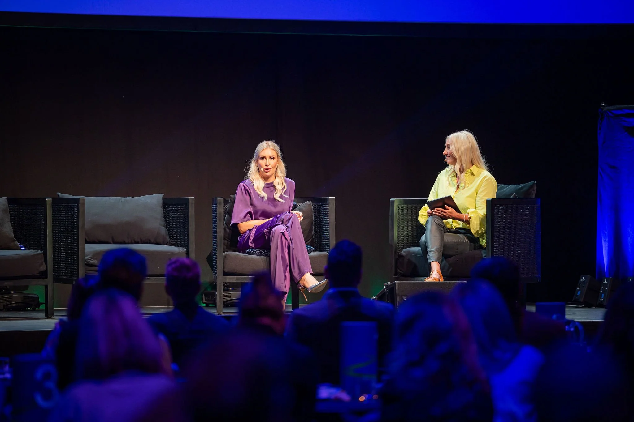 Two women seated on a stage, engaging in conversation during an event, with audience members visible in the foreground.
