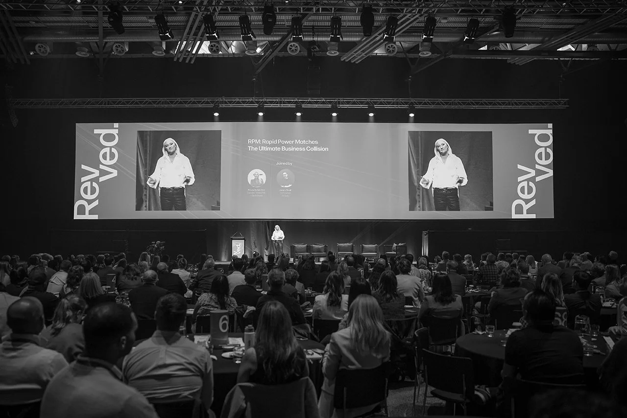 A black and white photo of a conference or seminar with a large audience watching a speaker on stage. The stage has a big screen behind the speaker displaying the presentation titled 'RPM: Rapid Power Matches The Ultimate Business Collision'. The room is dimly lit with spotlights on the stage and high ceilings with visible pipes and equipment.