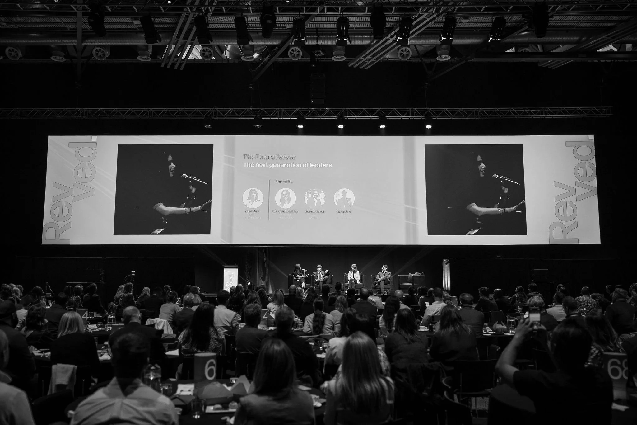 Black and white photo of a conference or panel discussion with five people sitting on stage, two women and three men, with a large audience in front. The background shows a screen with a woman speaking into a microphone, and the words "The Future For